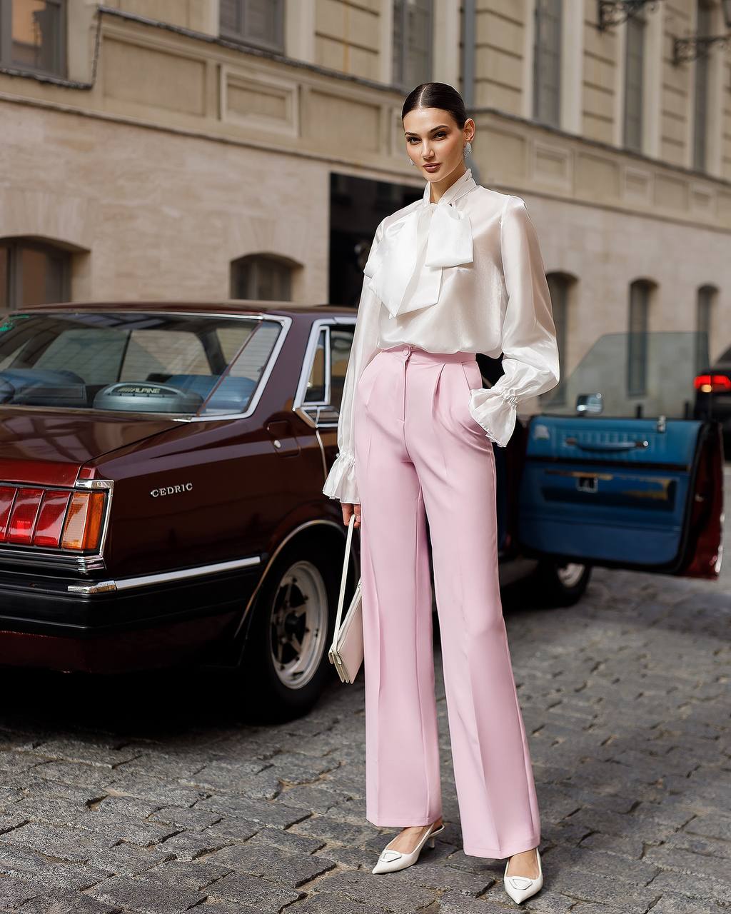 Woman in a white blouse and pink pants standing on a street with vintage cars in the background