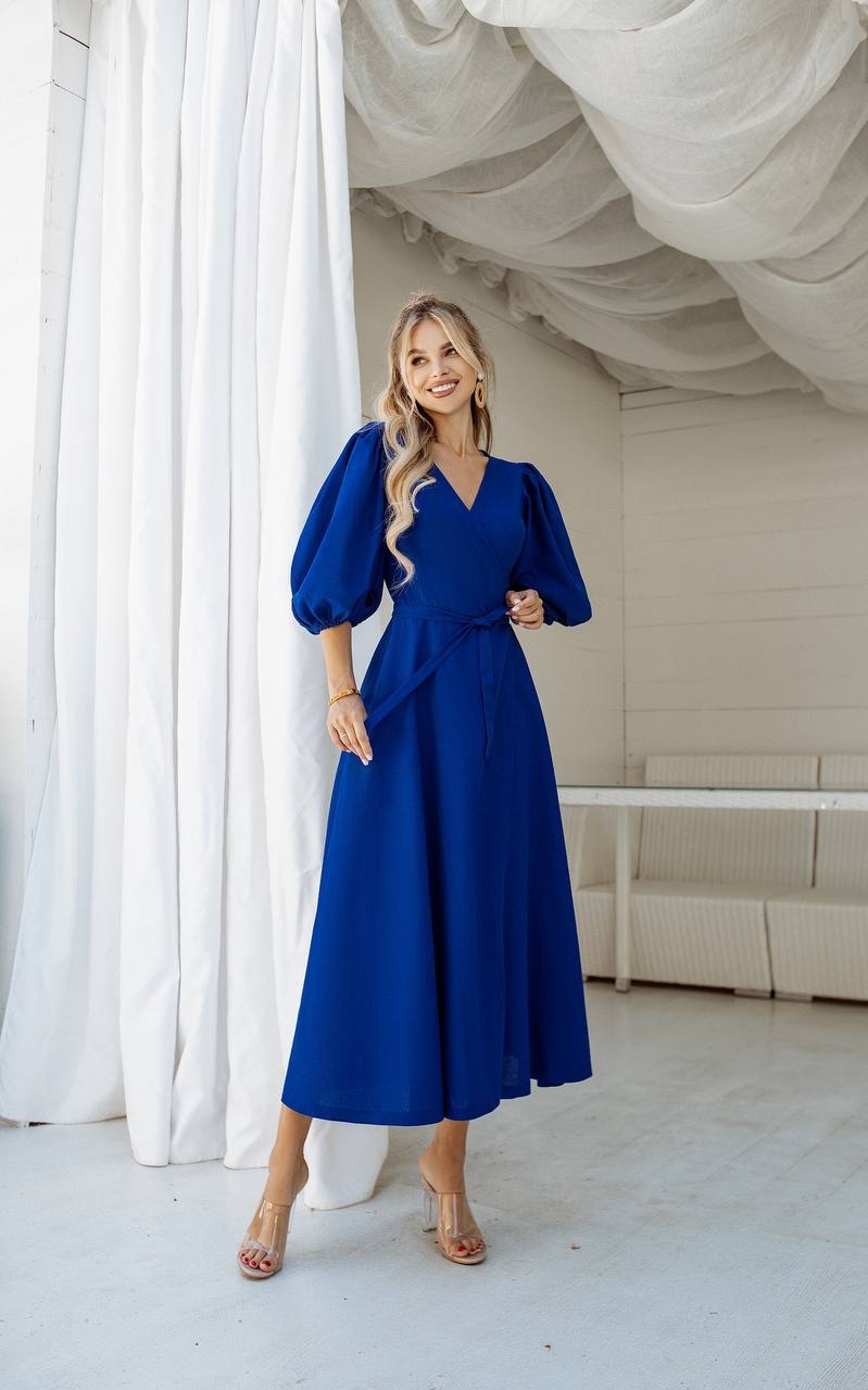 Woman in a blue dress standing in a room with white curtains and a beige sofa.