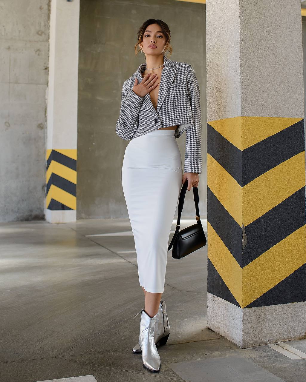Woman in a checkered blazer, white skirt, and silver boots standing in an urban setting with yellow and black striped barriers.