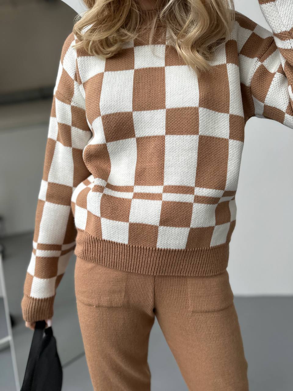 Person wearing a brown and white checkered sweater with beige pants indoors.