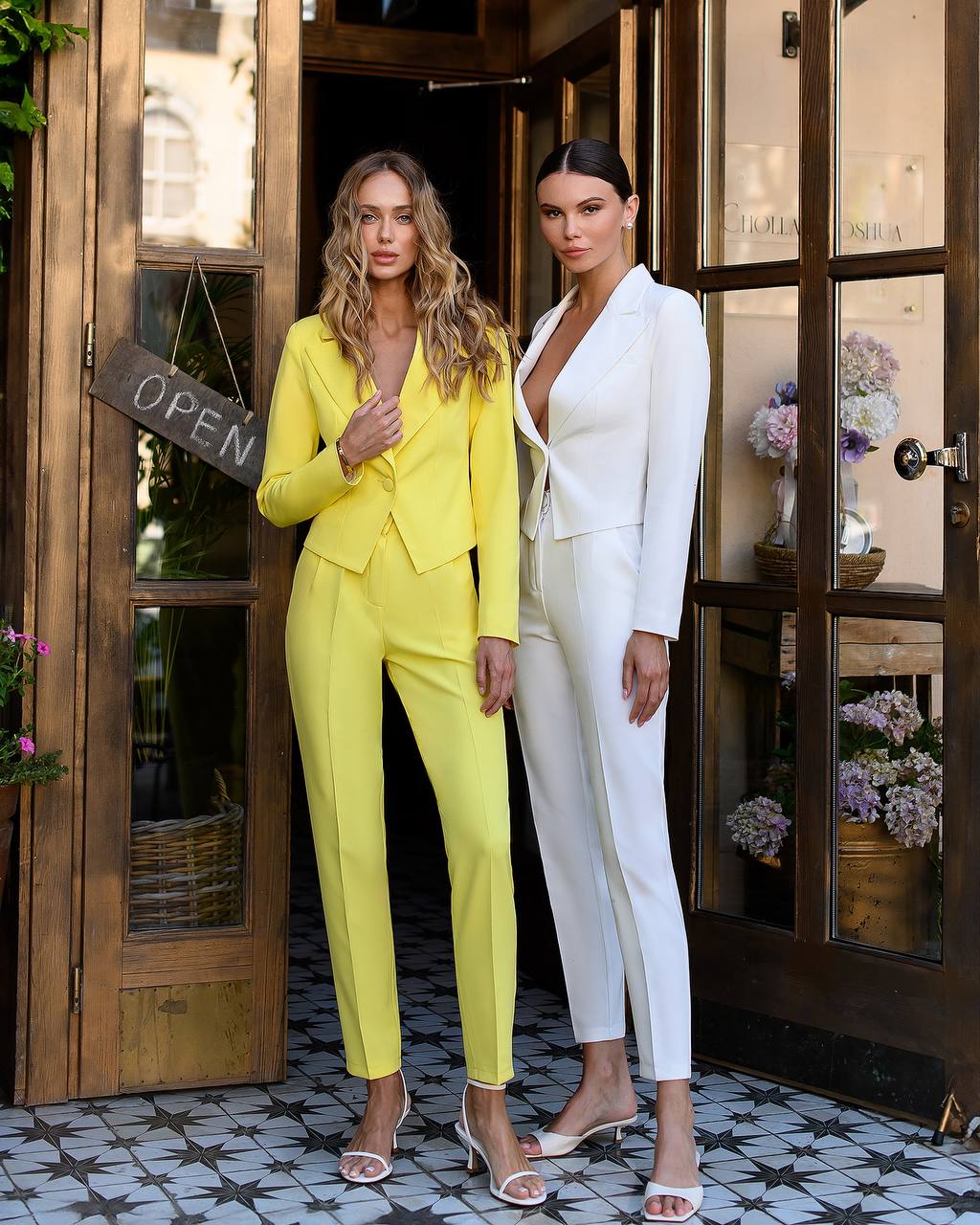 Two women in yellow and white suits standing in front of a door with 'Open' sign.