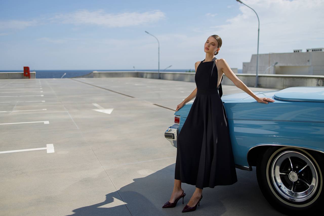 Woman in a black dress standing next to a blue vintage car on a parking lot.