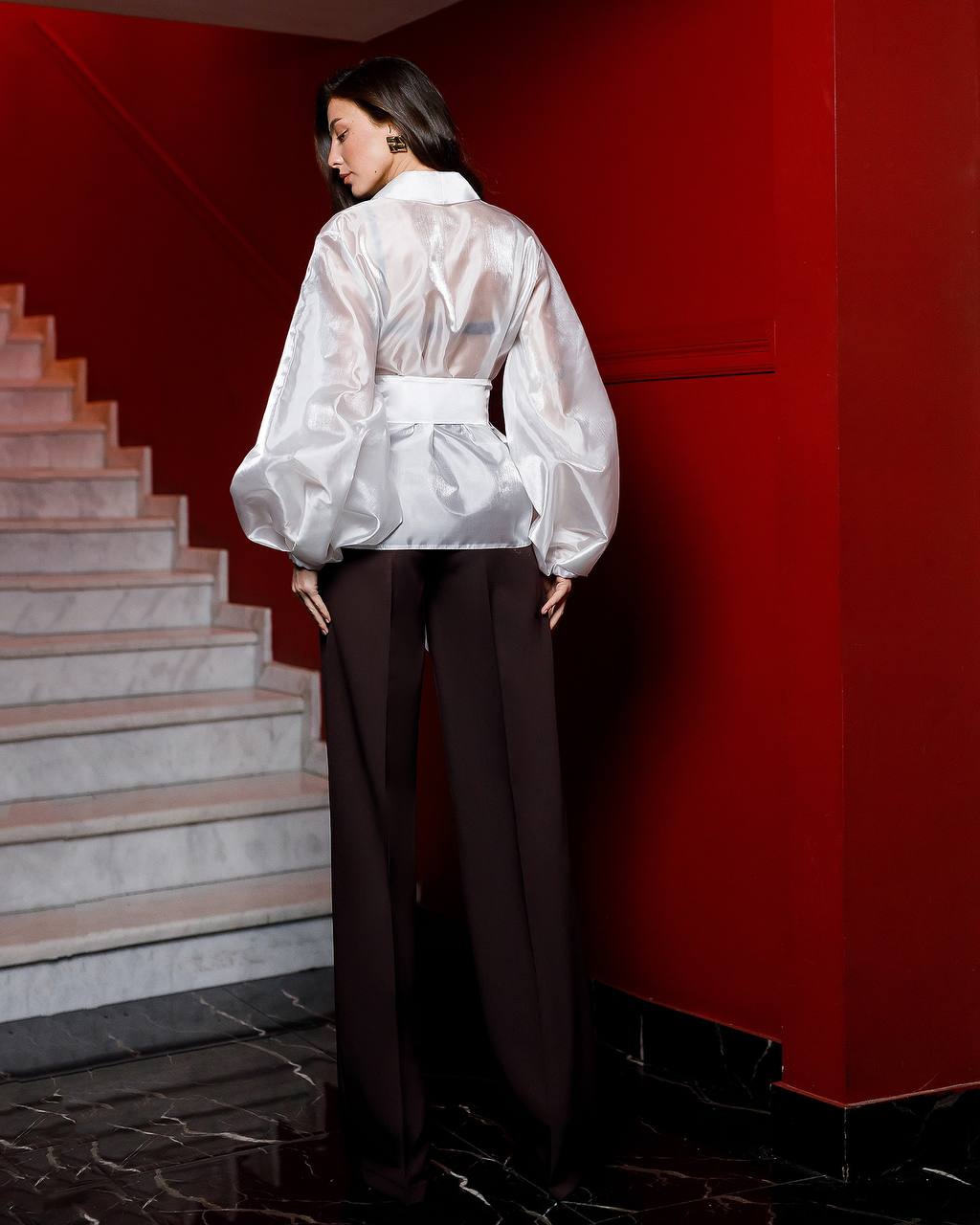 Person wearing a white sheer blouse and dark pants against a red wall.