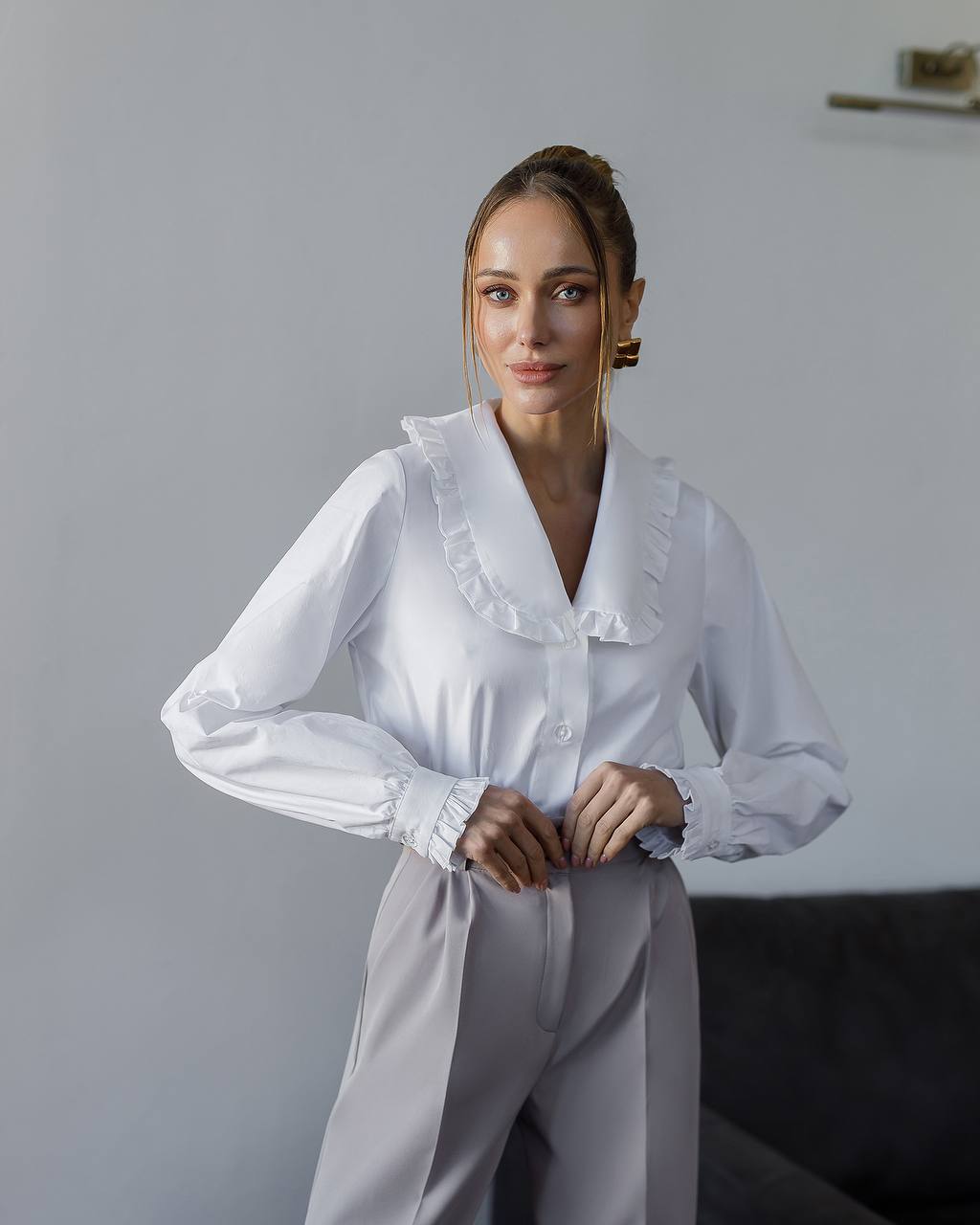 Woman wearing a white blouse and light gray pants against a plain background