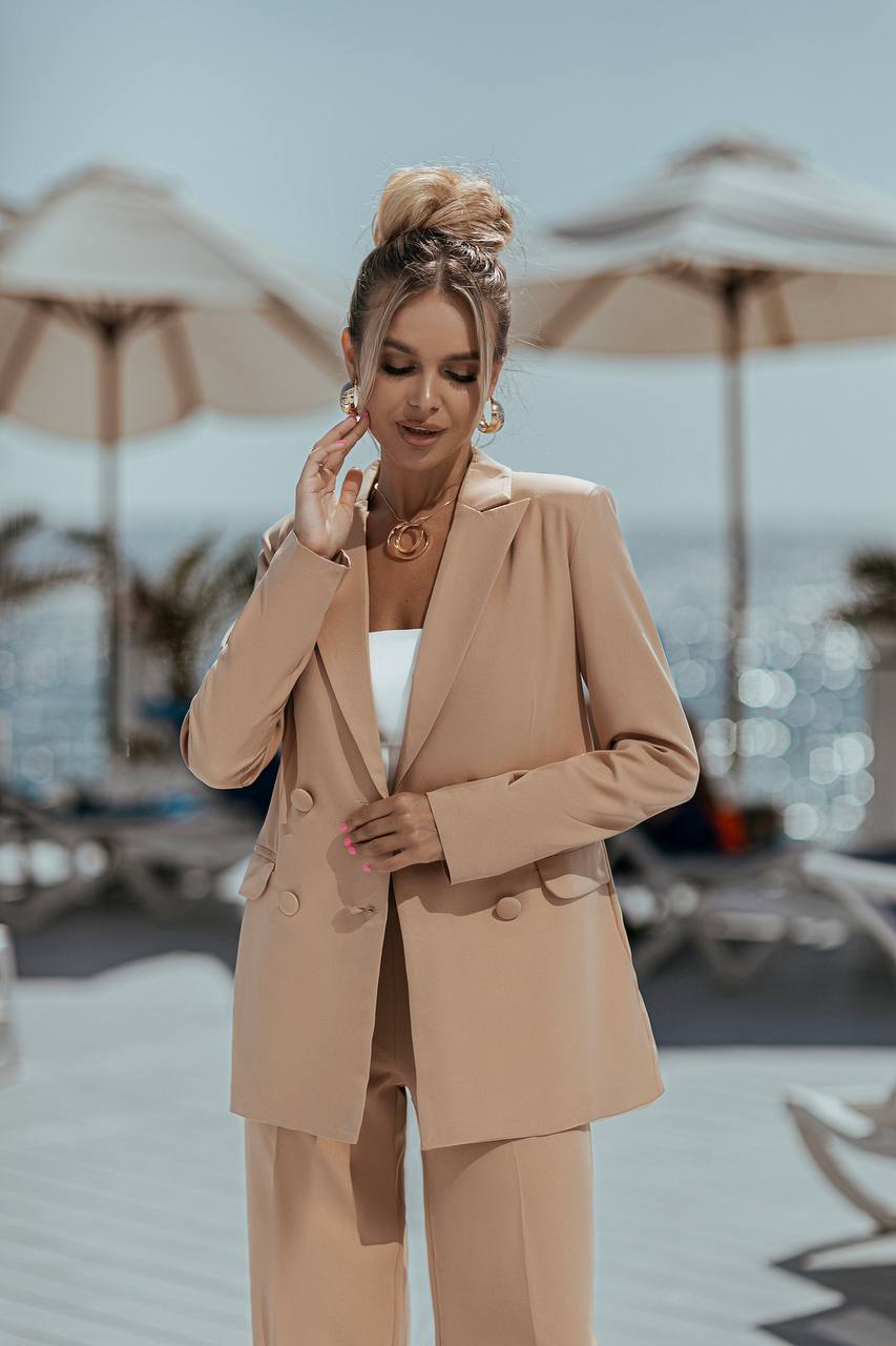 Woman in a beige suit standing by a poolside with umbrellas and lounge chairs.