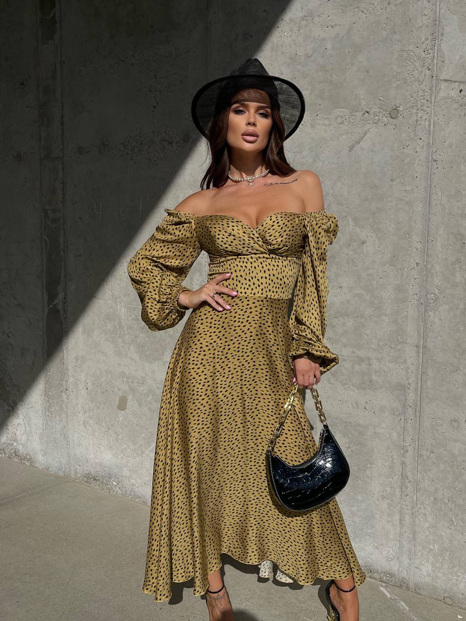 Woman in a patterned dress and black hat standing against a concrete wall.