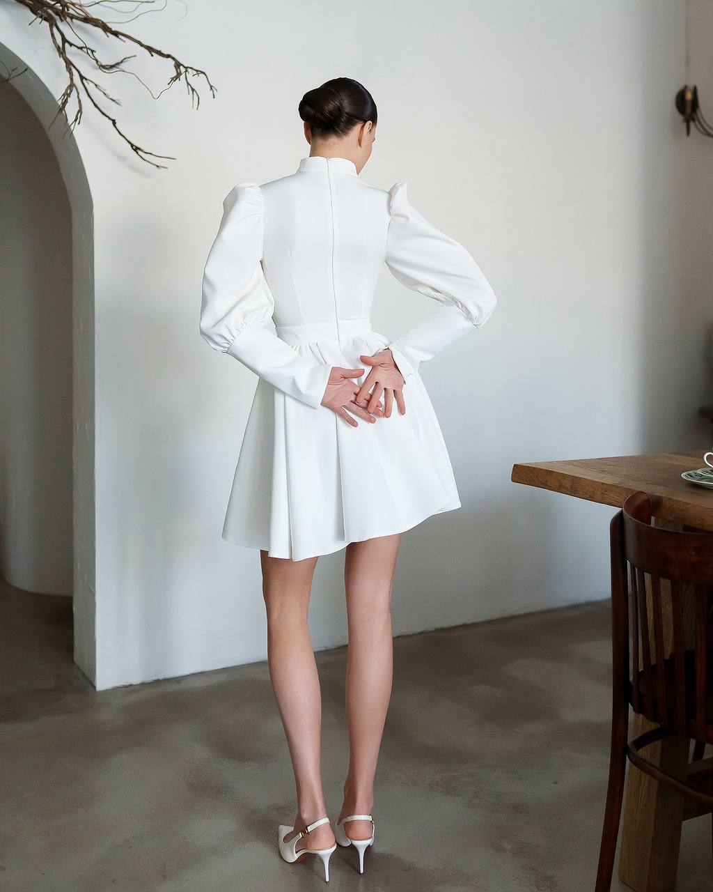 Woman in a white dress standing in a minimalistic room with a wooden table and chair.