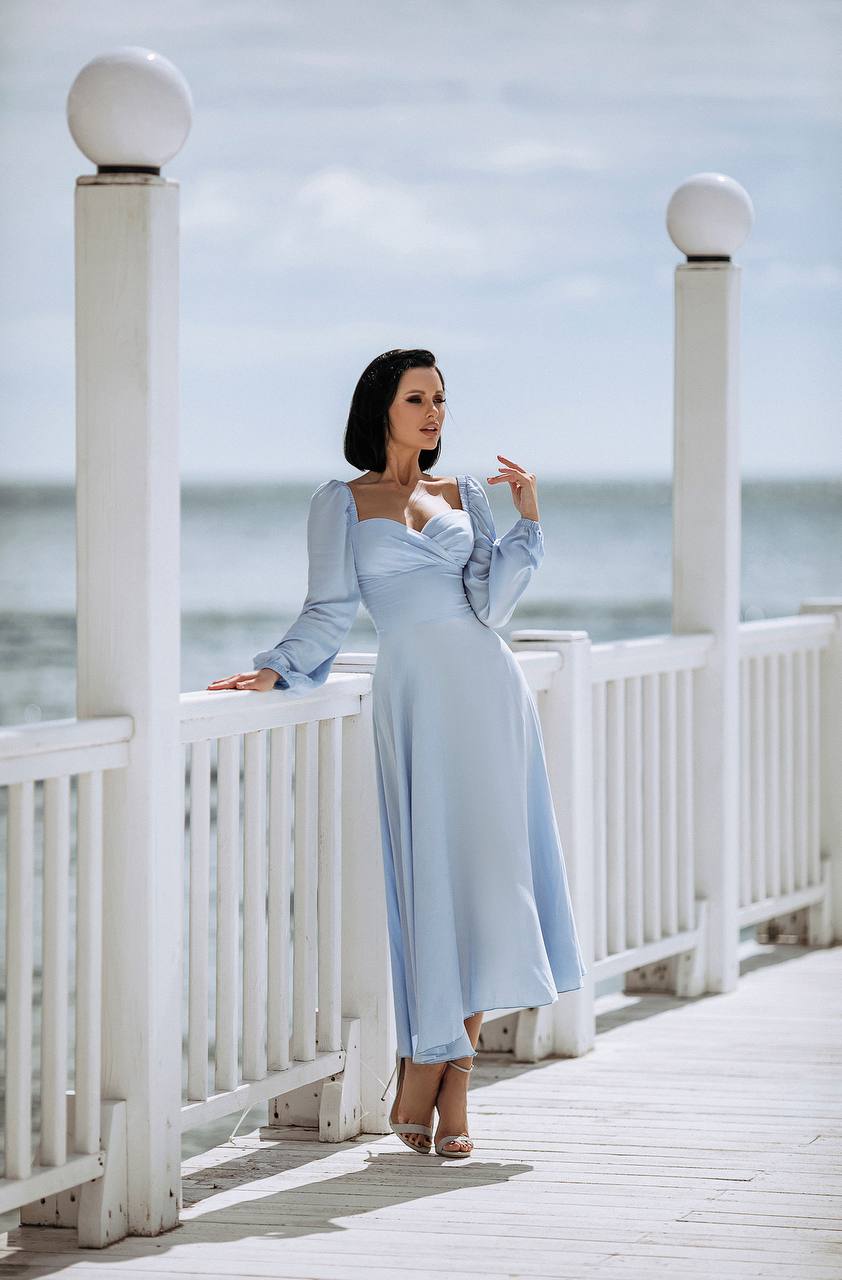 Woman in a light blue dress standing on a white wooden deck by the sea.