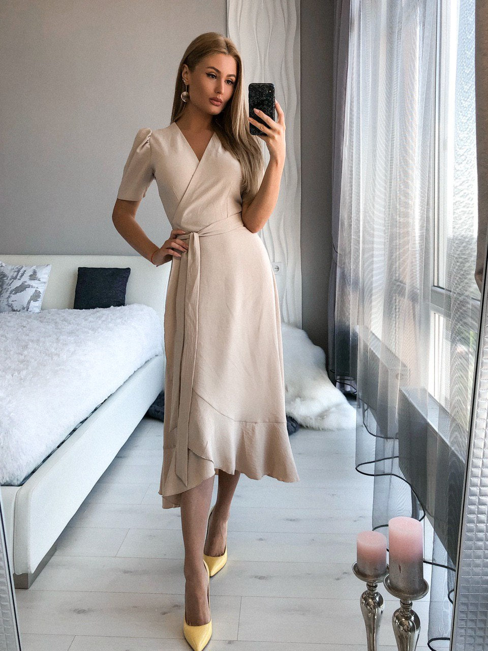 Woman taking a mirror selfie wearing a beige wrap dress in a modern room.