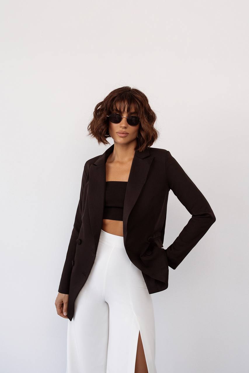 Woman wearing a black blazer, black top, and white pants against a light gray background