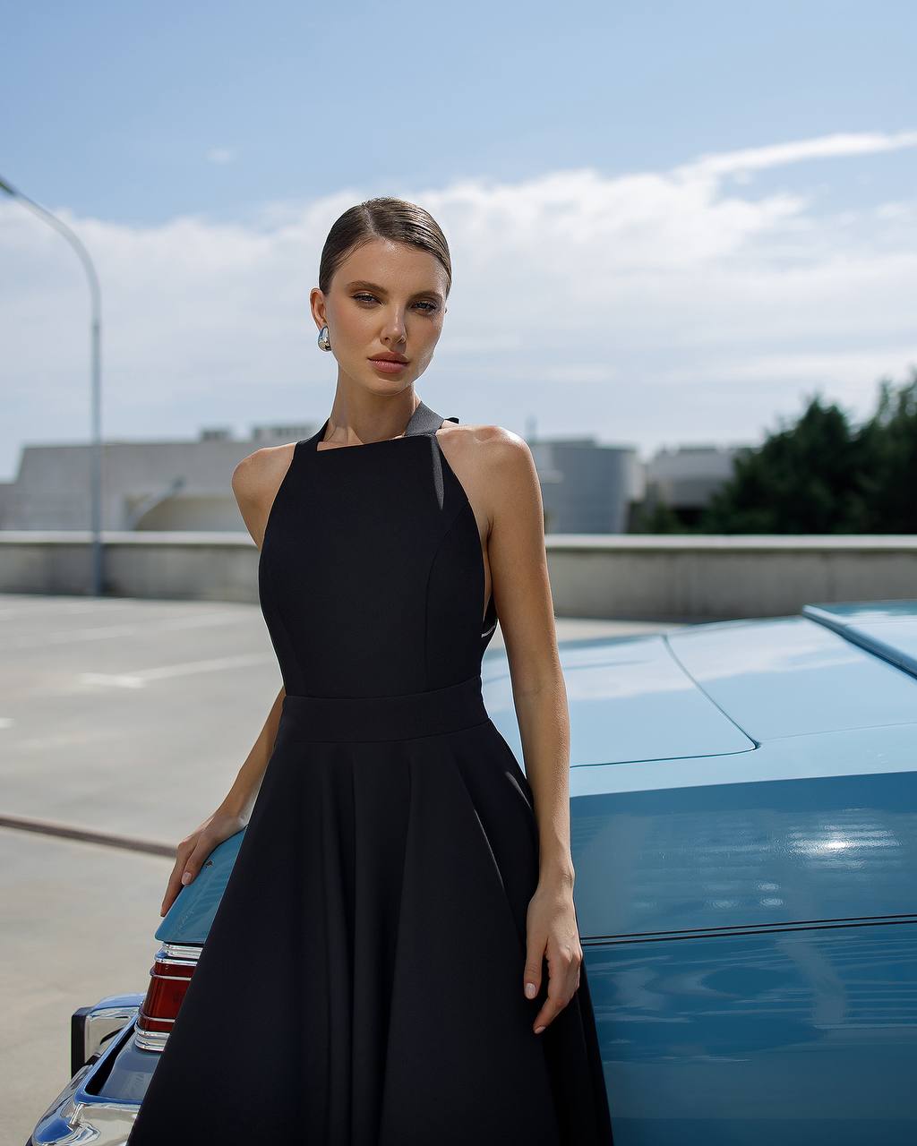 Woman in a black dress standing next to a blue car with a cityscape in the background