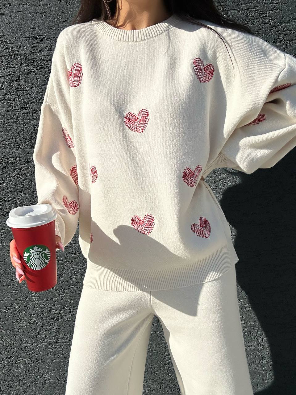 Person wearing a white sweater with red heart patterns, holding a red Starbucks cup against a dark background.