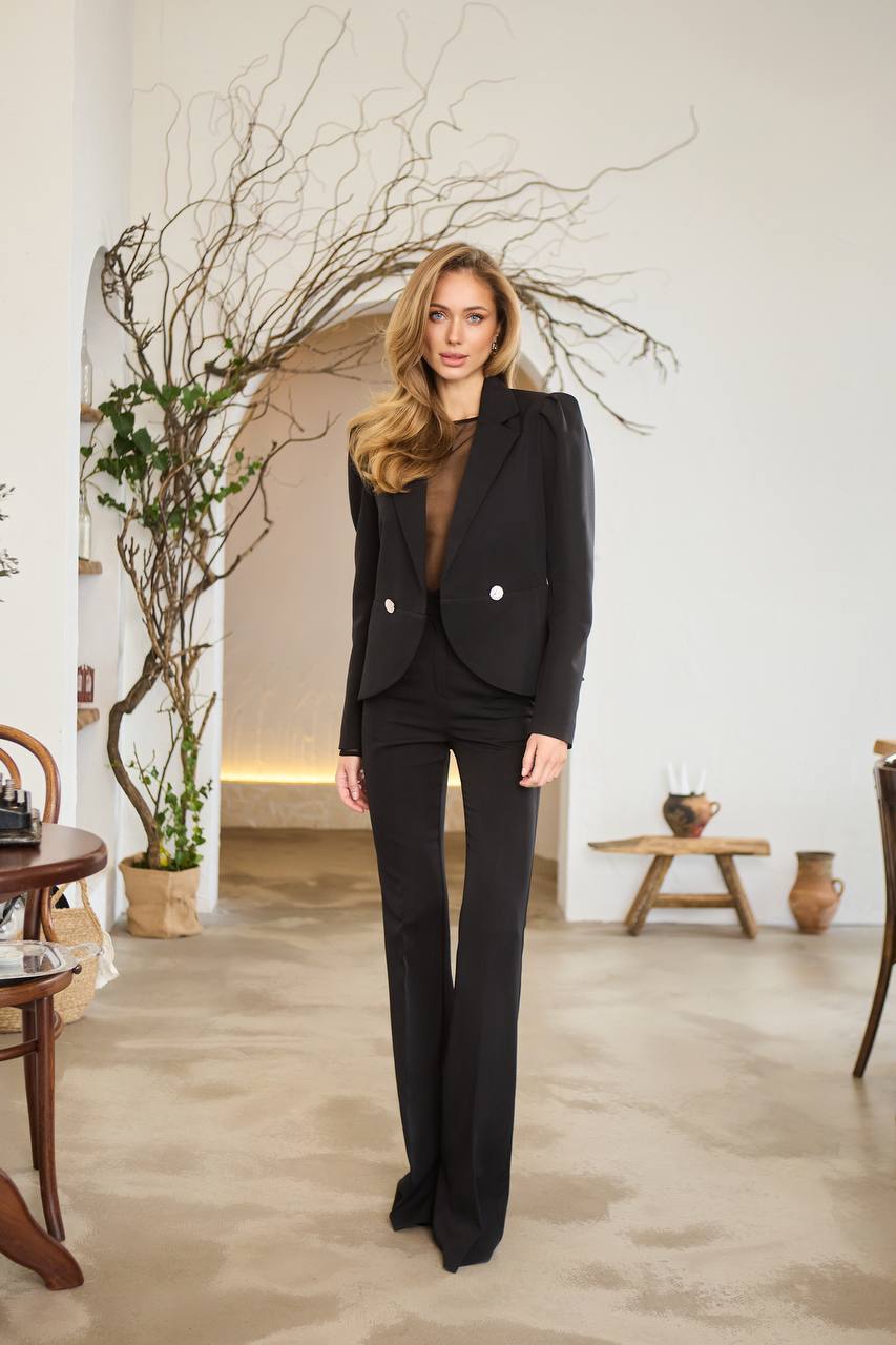 Woman in a black suit standing in an indoor setting with decor elements.