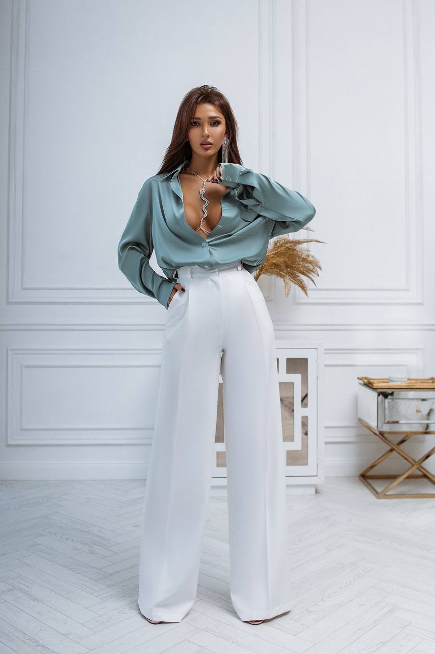 Woman wearing a light blue blouse and white pants in a room with white walls and wooden floor.