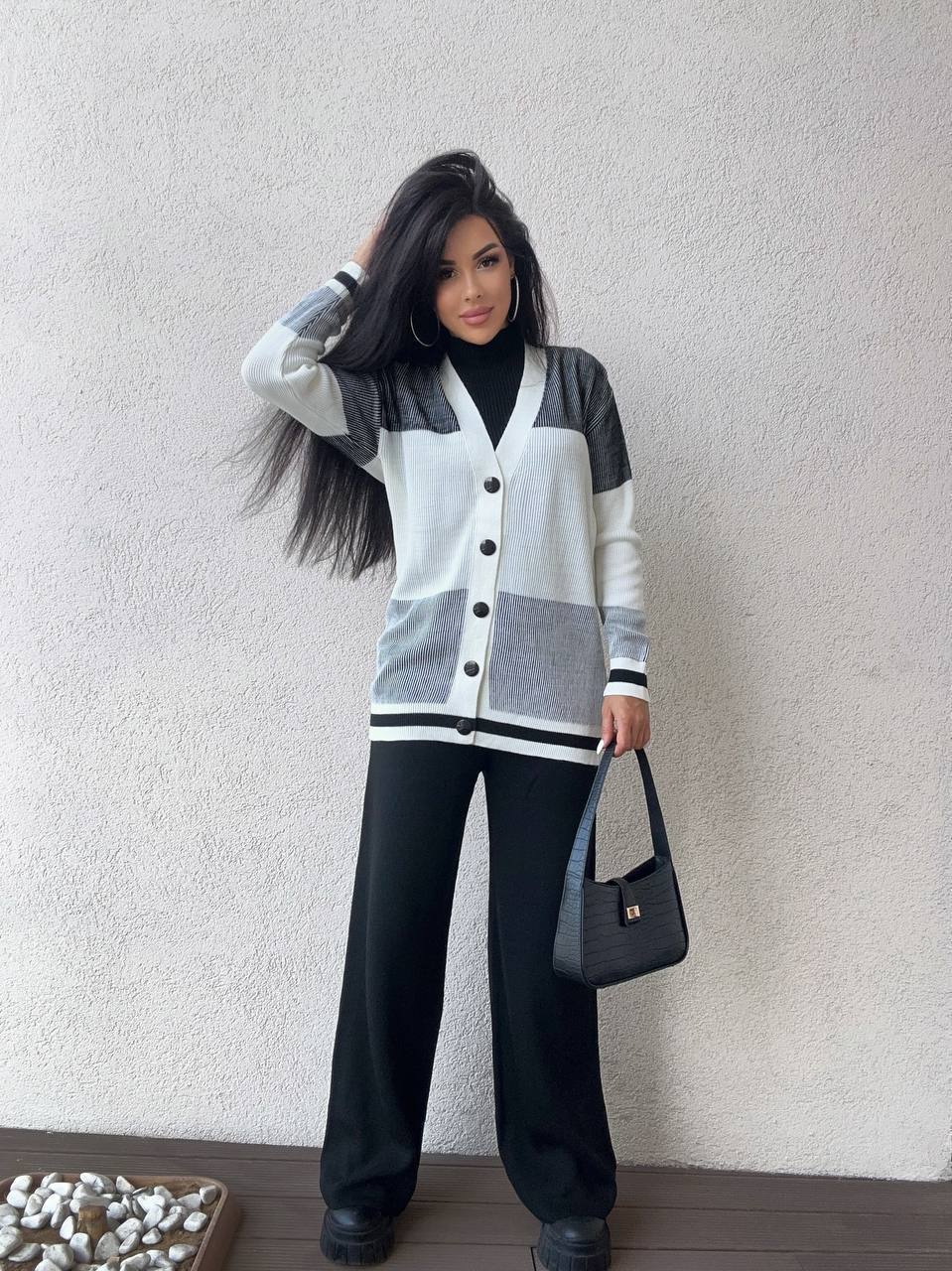 Woman wearing a gray and black cardigan with black pants against a plain wall.