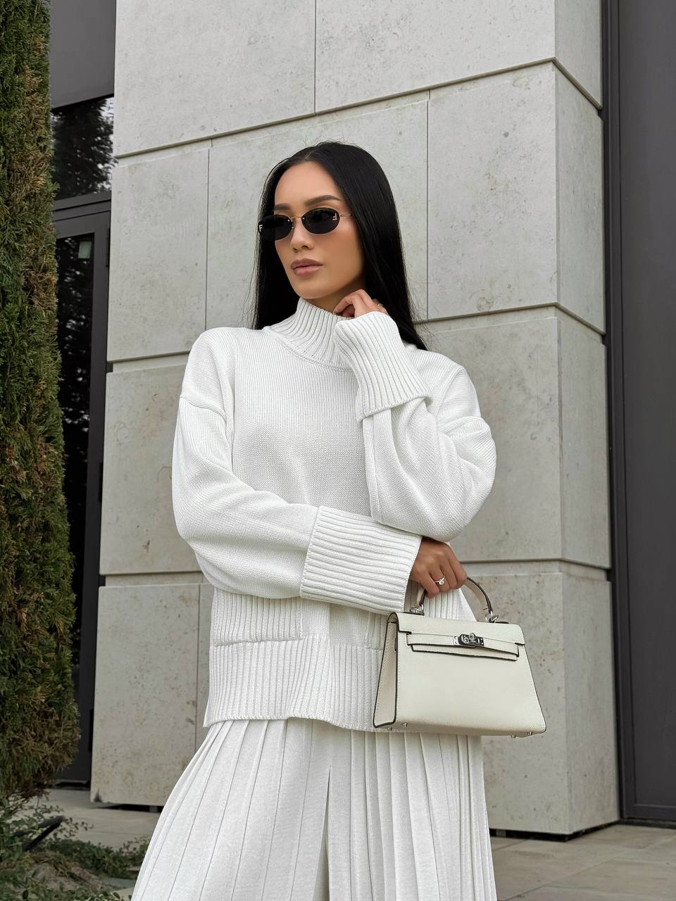 Woman in a white outfit holding a cream handbag against a light stone wall.