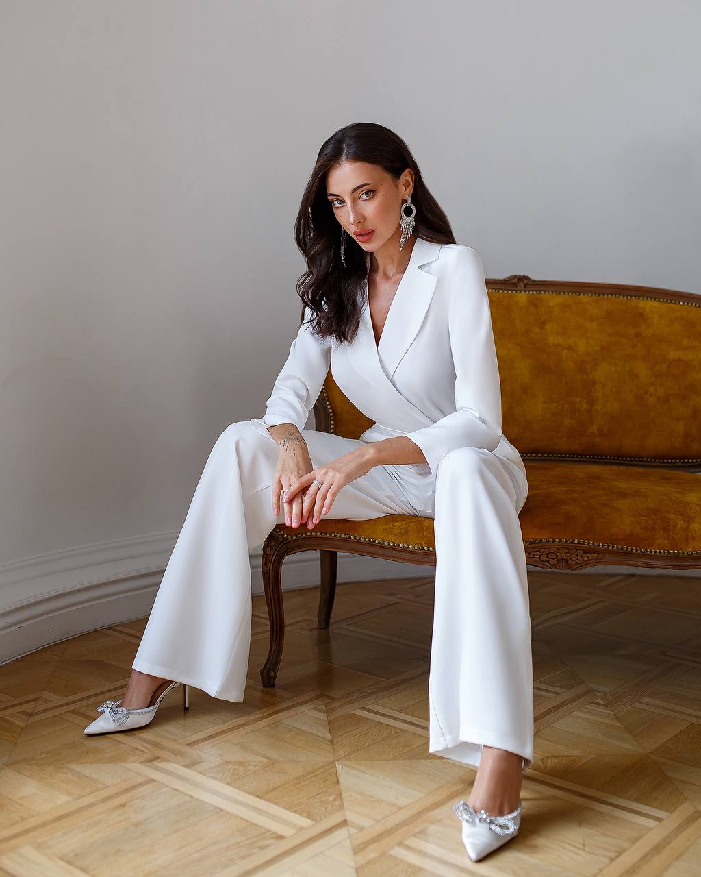 Woman in a white suit sitting on a vintage couch in a room with wooden flooring.