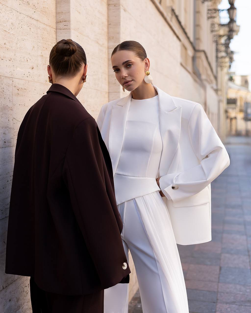 Two models standing on a city street. One is wearing a structured white suit with a matching top, and the other is wearing a dark brown blazer. They are posed against a light-colored stone building wall.