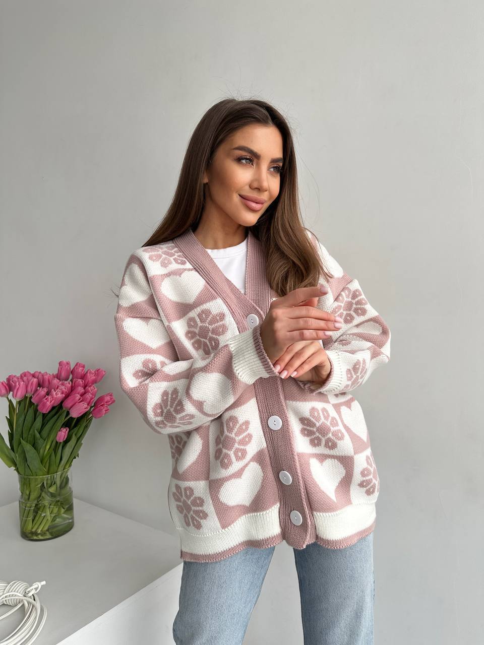 Woman wearing a patterned cardigan with a vase of pink tulips on a white background