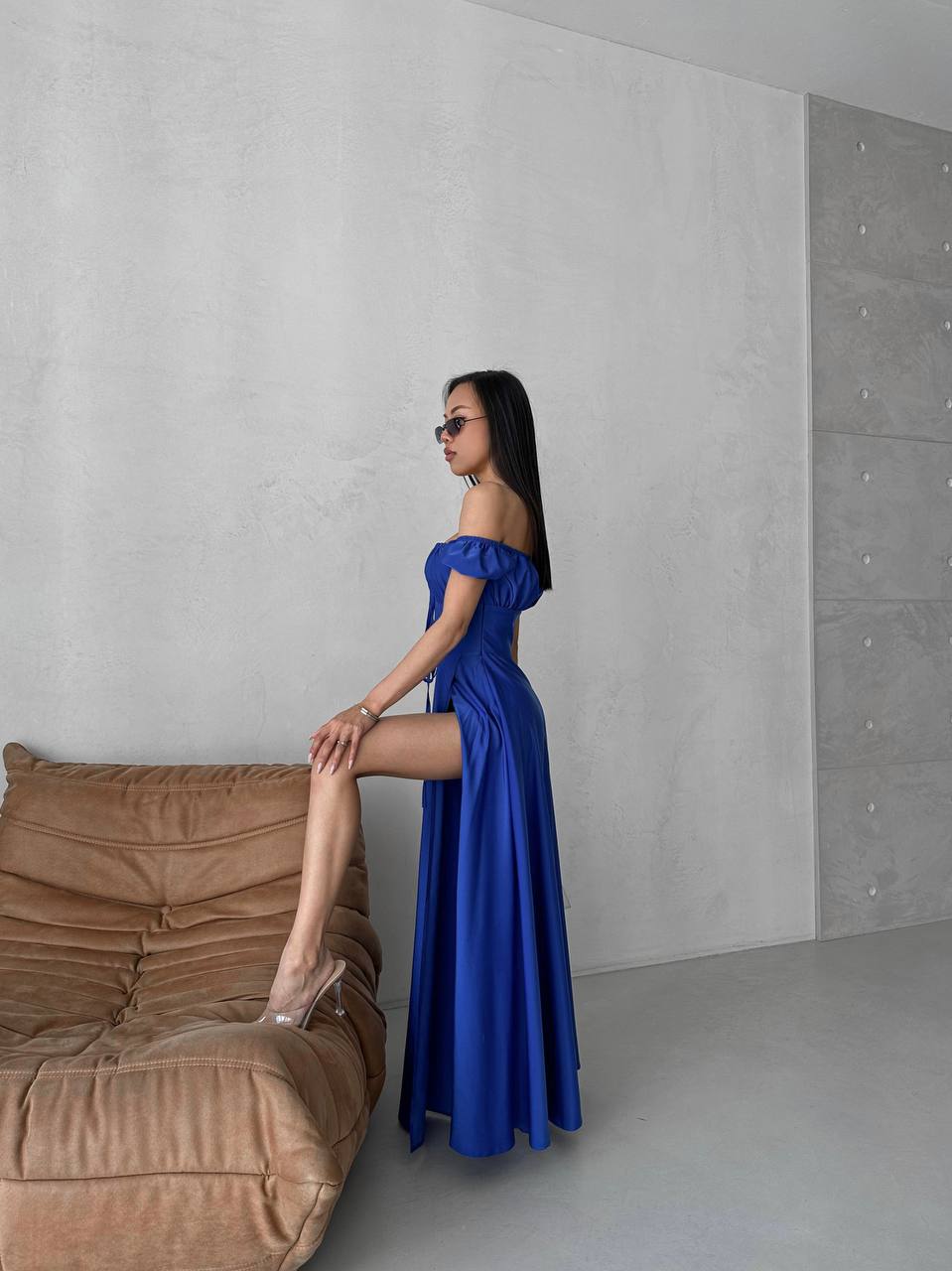 Woman in a blue dress posing next to a brown sofa against a gray wall.