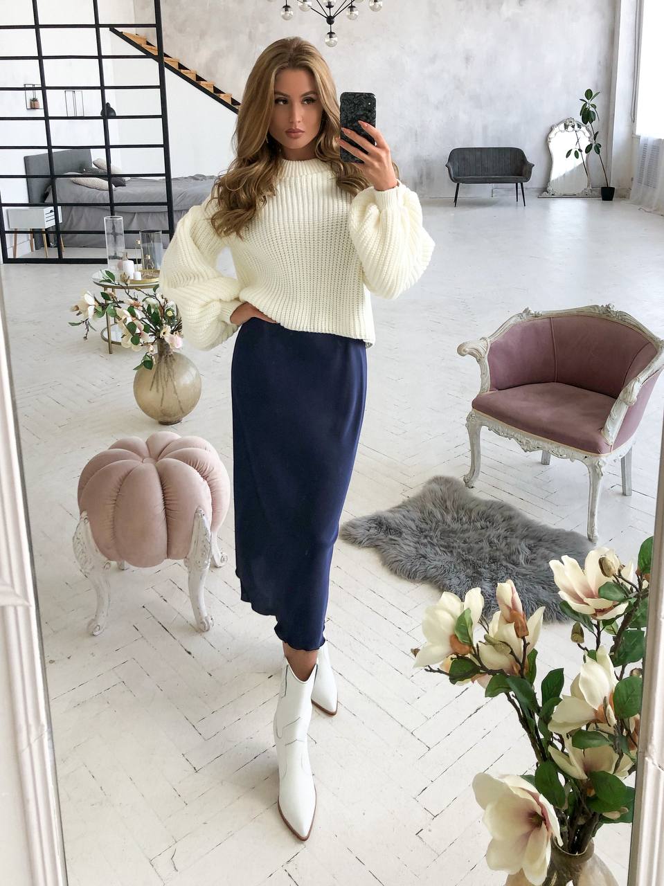 Woman taking a mirror selfie wearing a white sweater and navy skirt in a stylish room.