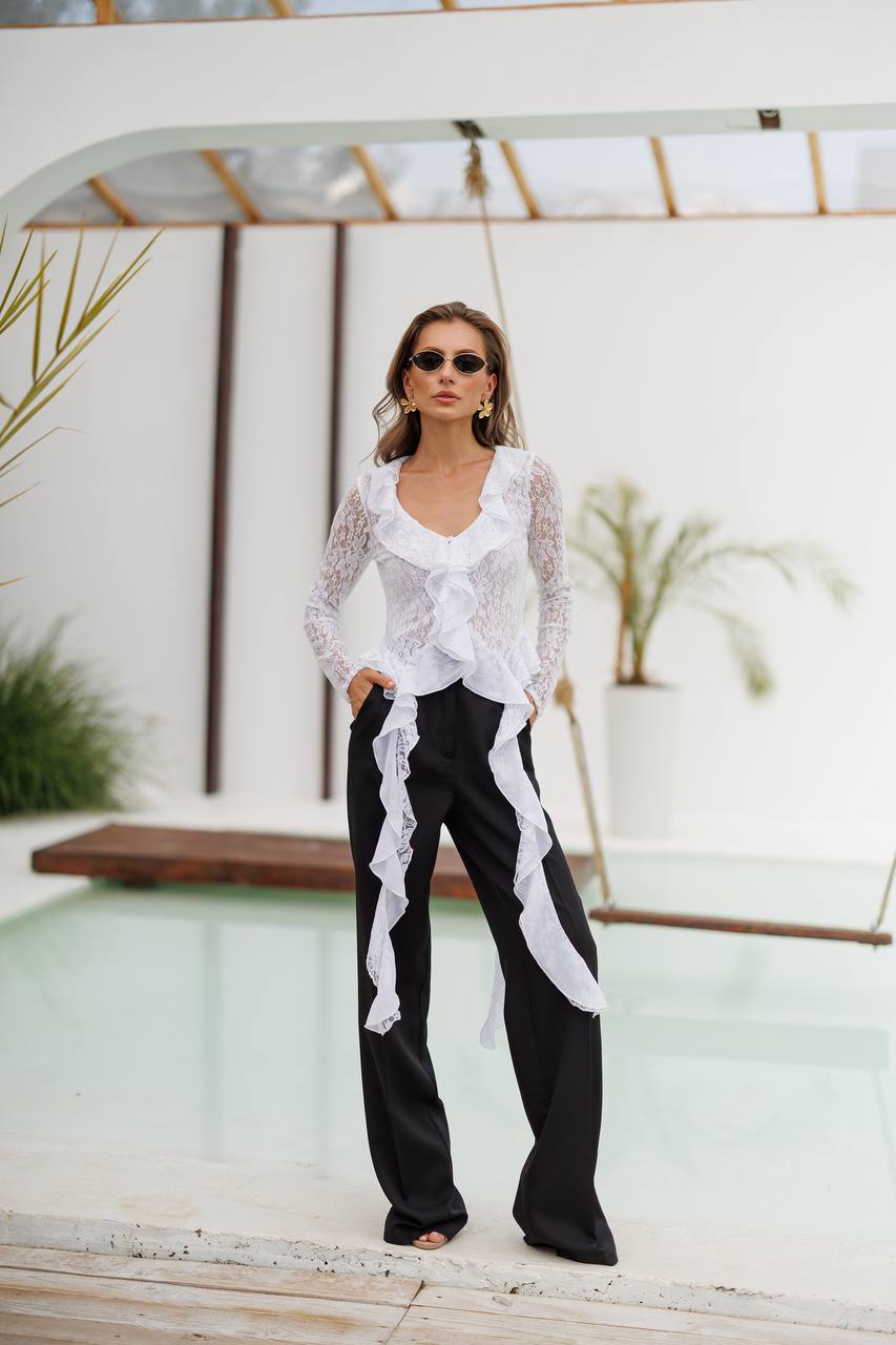Woman wearing a white lace top and black pants by a poolside.