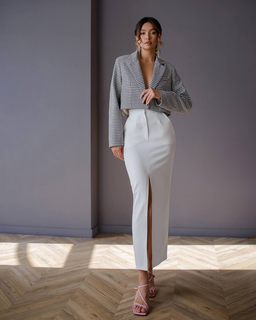 Woman in a checkered blazer and white pants standing in a room with a gray wall and wooden floor.