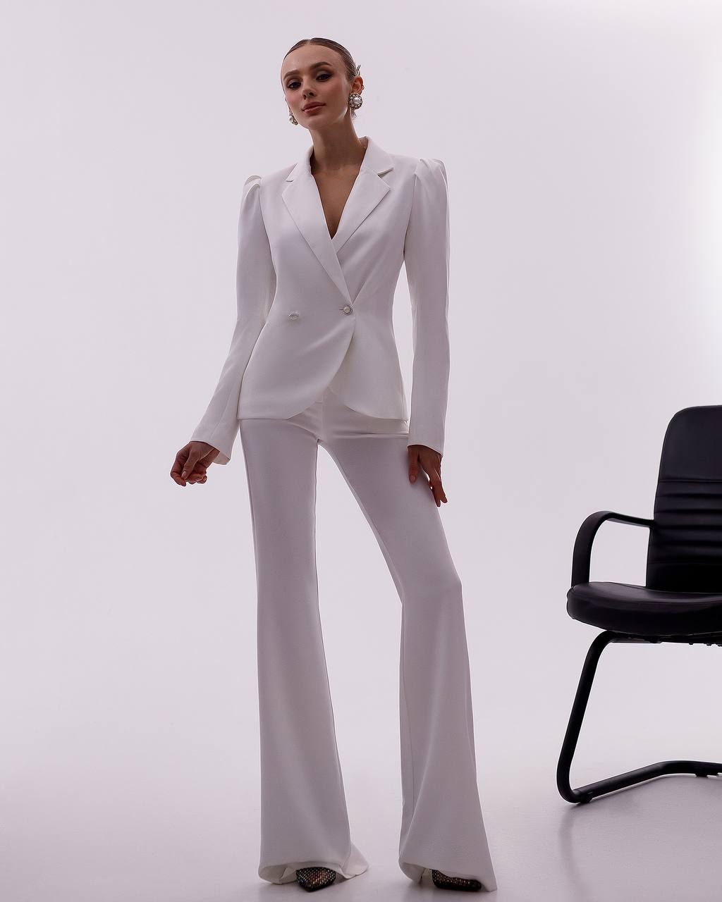 Woman wearing a white suit standing next to a black office chair on a white background