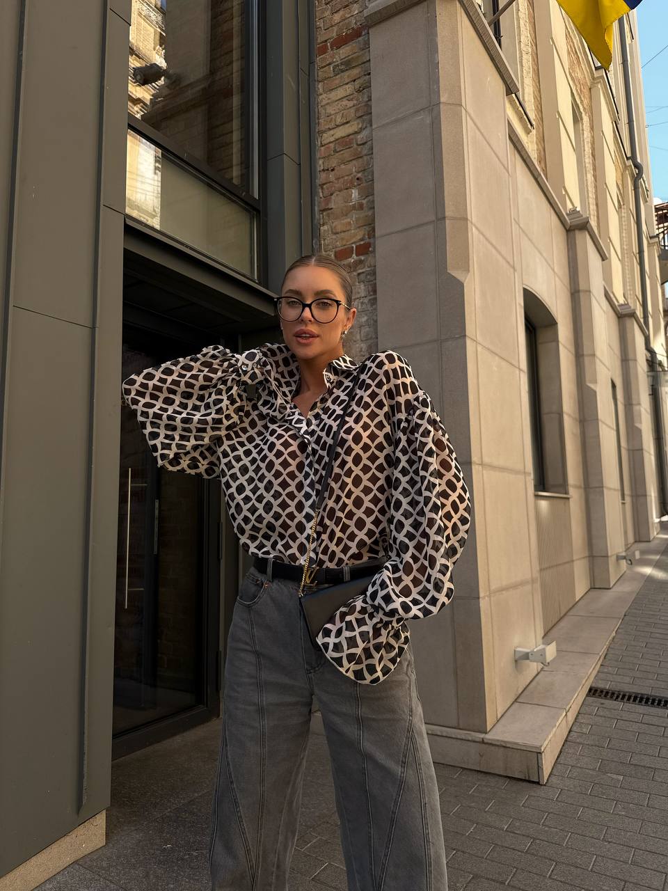 Person wearing a patterned shirt and jeans standing on a street with buildings in the background