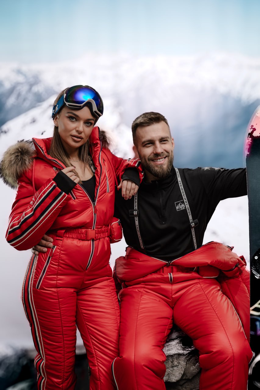 Two people in red ski suits posing on a mountain with snow and clouds in the background.