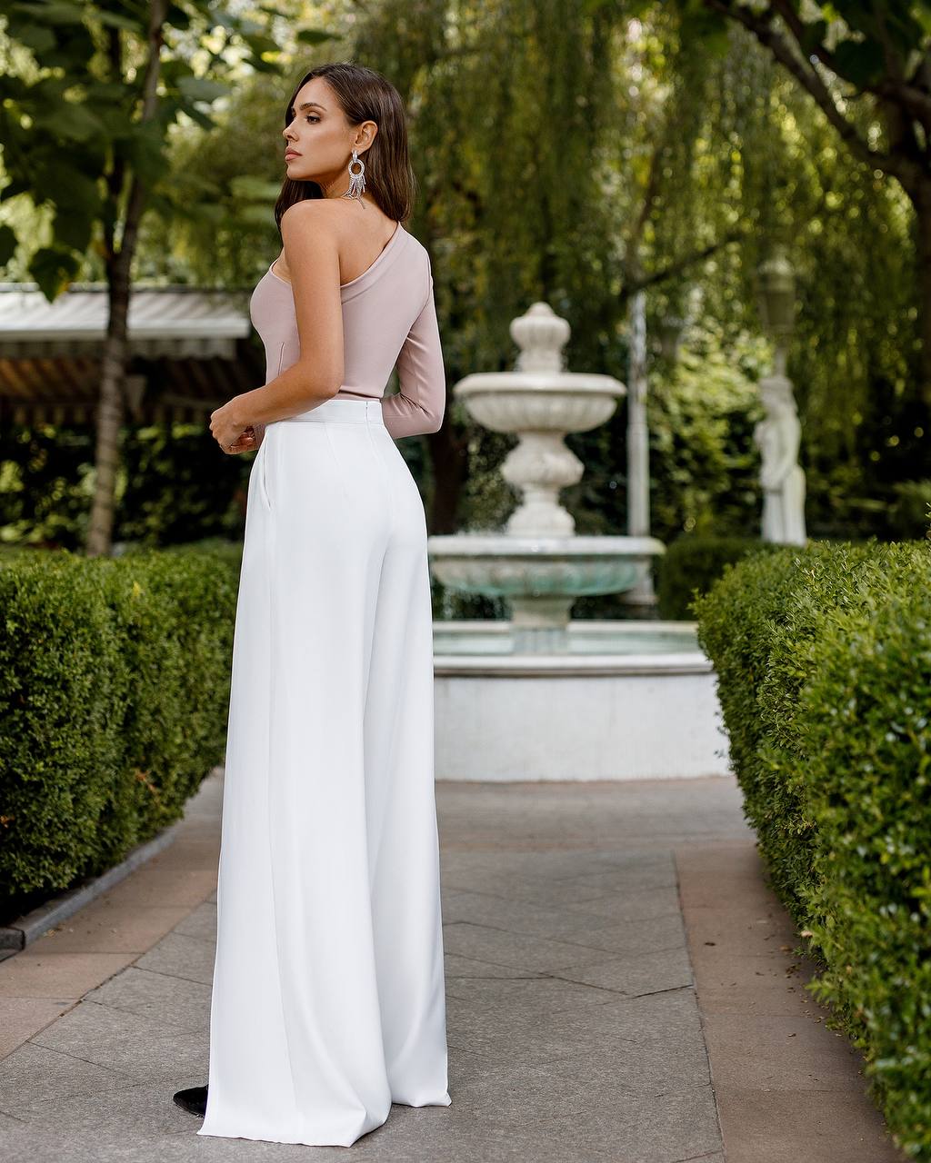 Woman in a pink top and white pants standing in a garden with a fountain.