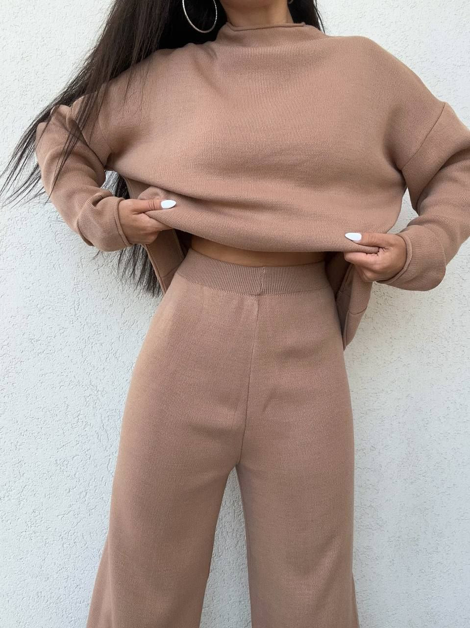 Person wearing a brown cropped sweater and high-waisted pants against a white wall