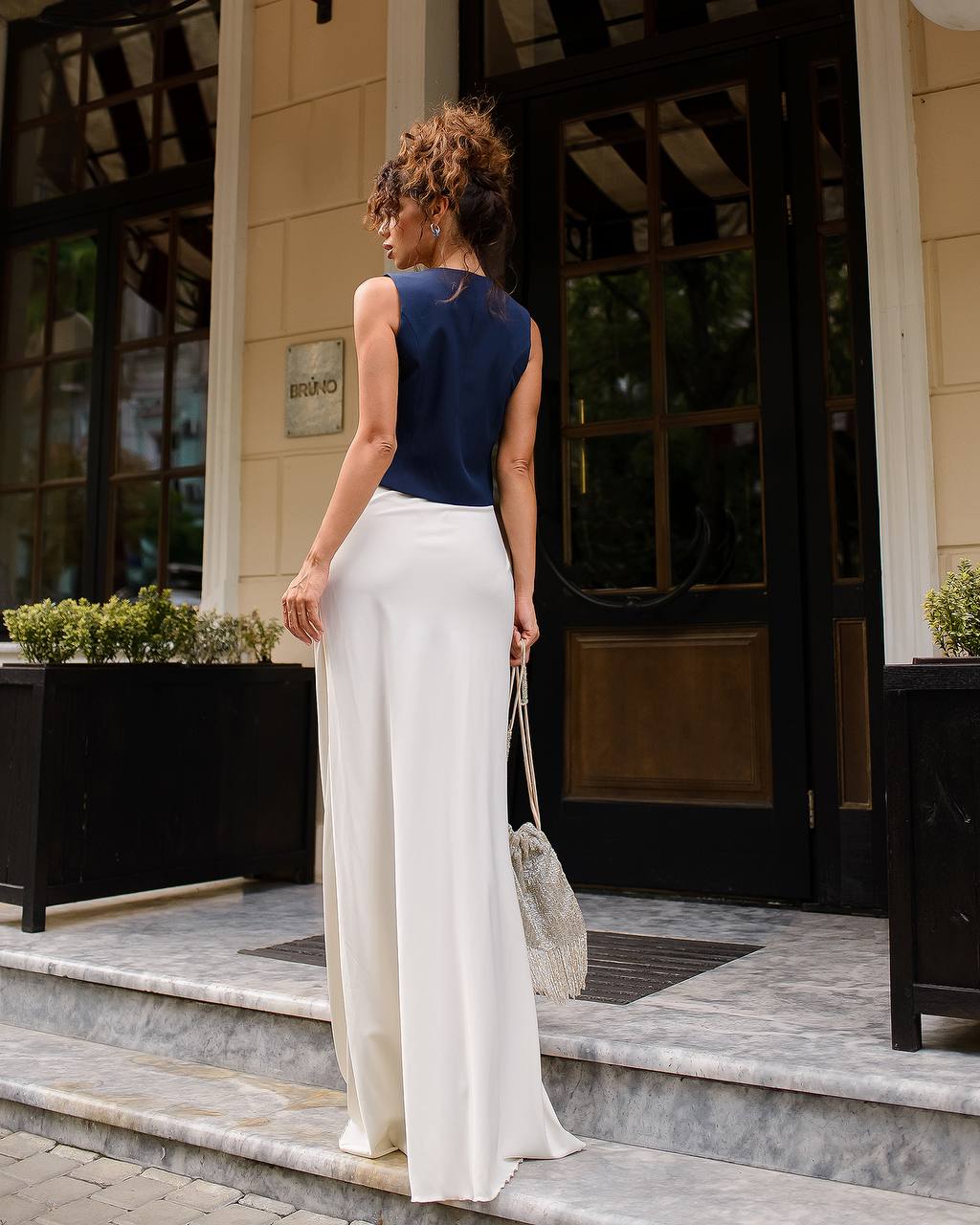 Woman in a blue top and white pants standing on a sidewalk.