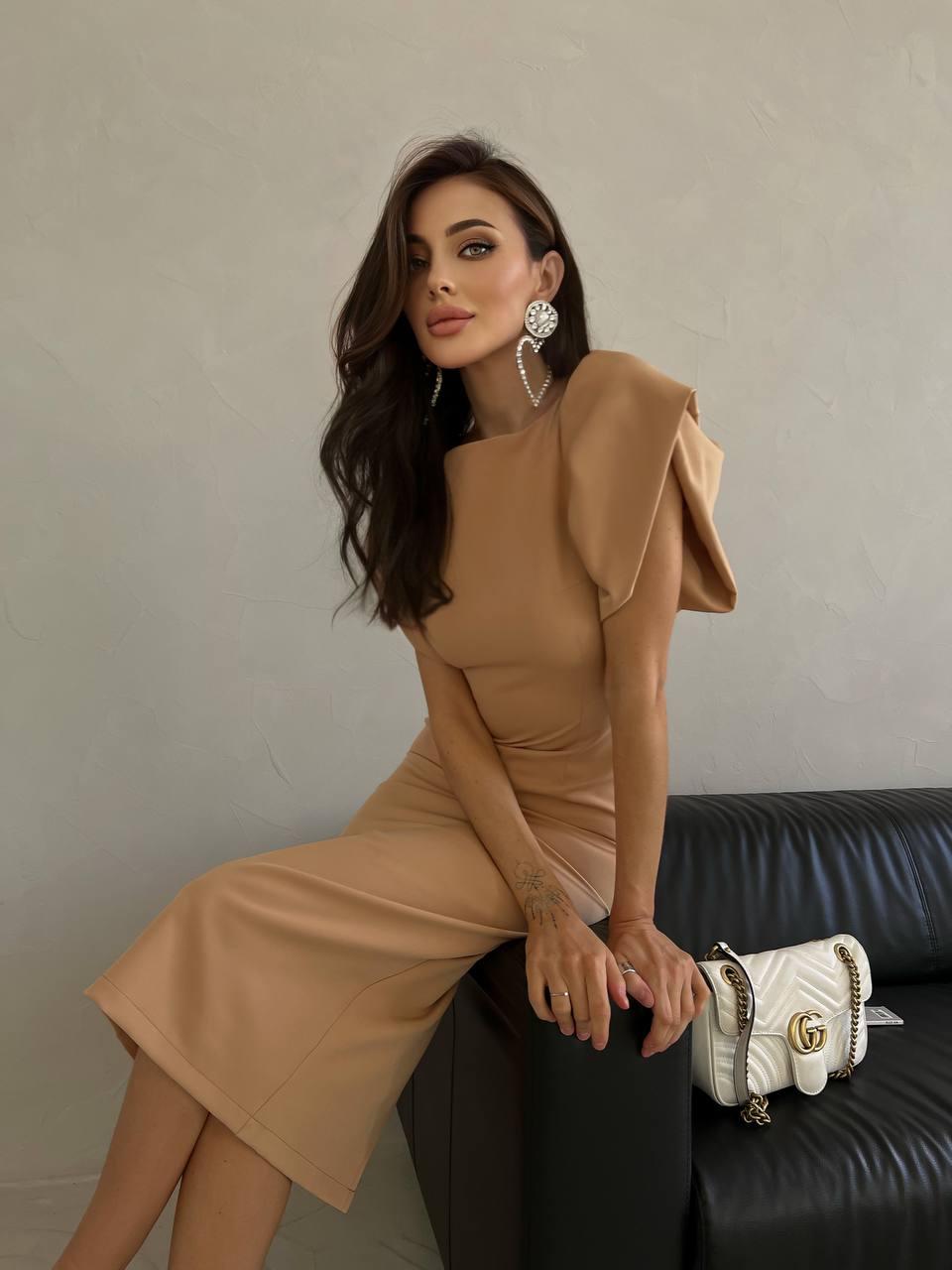 Woman in a beige dress sitting on a black couch with a white handbag.