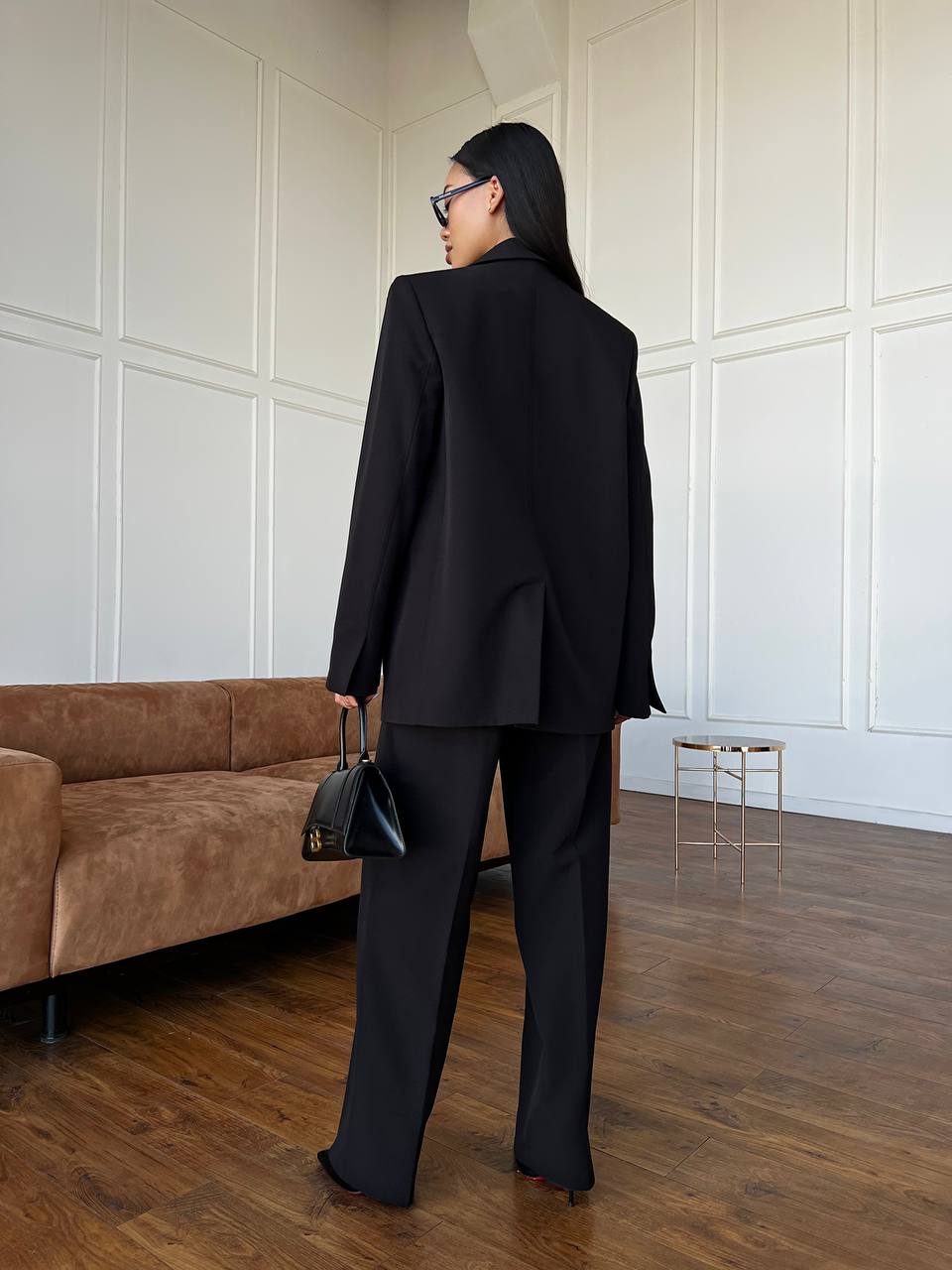 Person wearing a black suit standing in a room with a brown couch and white paneled walls.