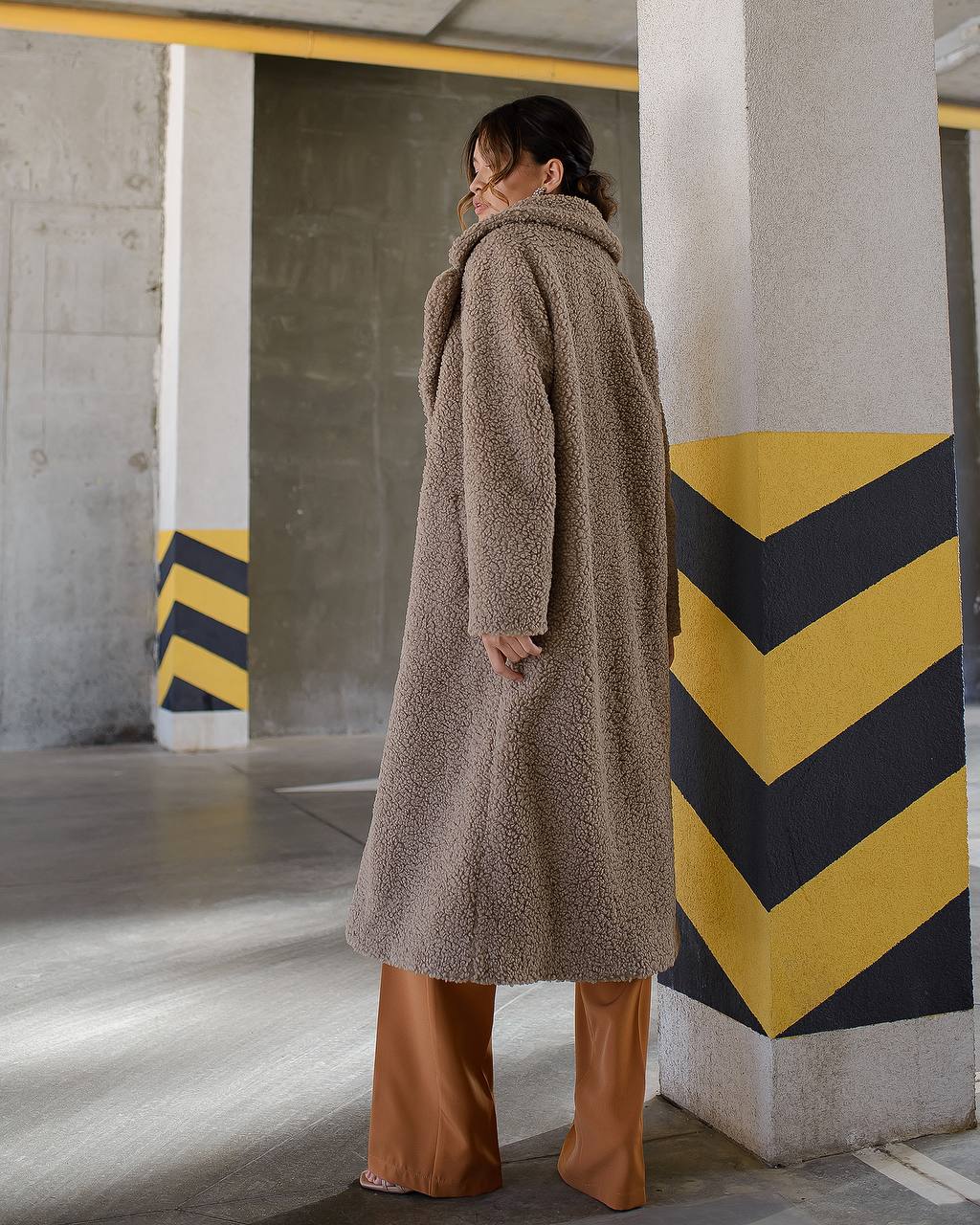 Person wearing a long brown coat in an urban setting with concrete pillars and yellow and black stripes.