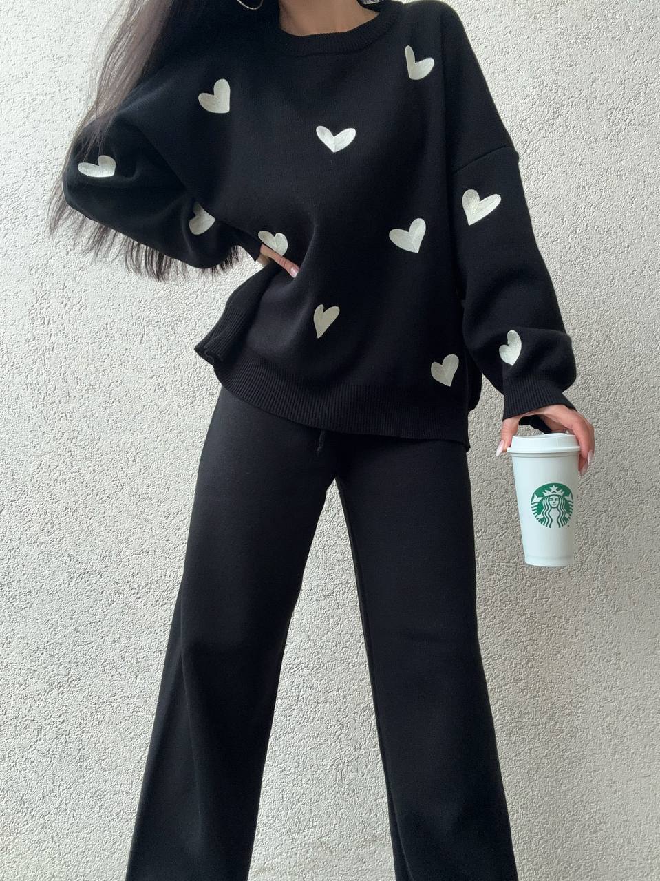 Person wearing a black sweater with heart patterns and holding a Starbucks cup against a light gray background