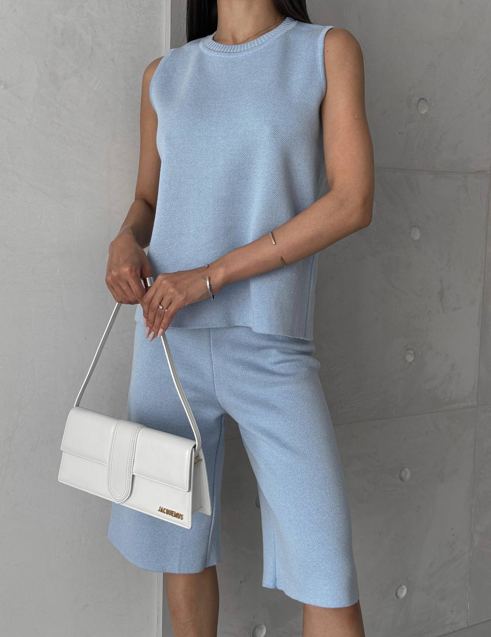 Person wearing a light blue sleeveless top and shorts holding a white handbag against a gray wall.