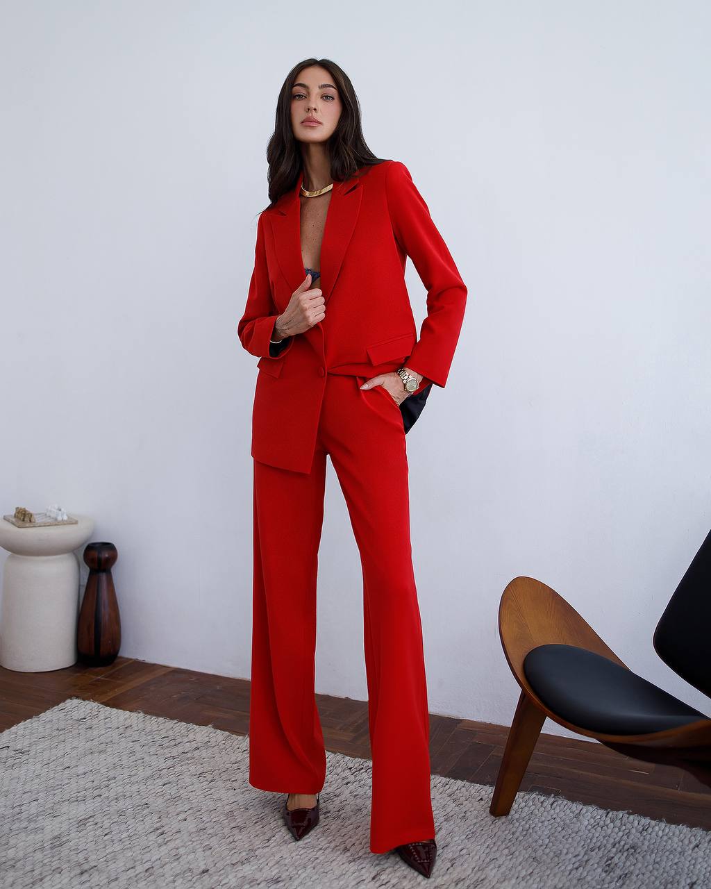 Woman wearing a red suit in a room with a chair and decorative items.