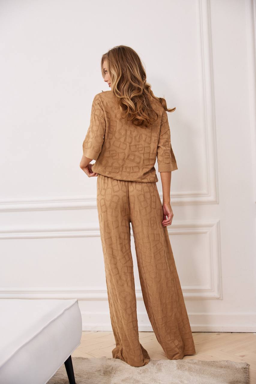 Woman wearing a brown textured outfit standing against a white wall.