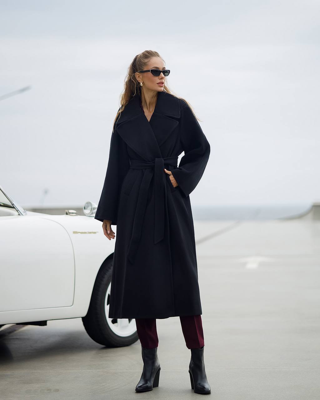 Woman in a long black coat standing next to a white car on a road.