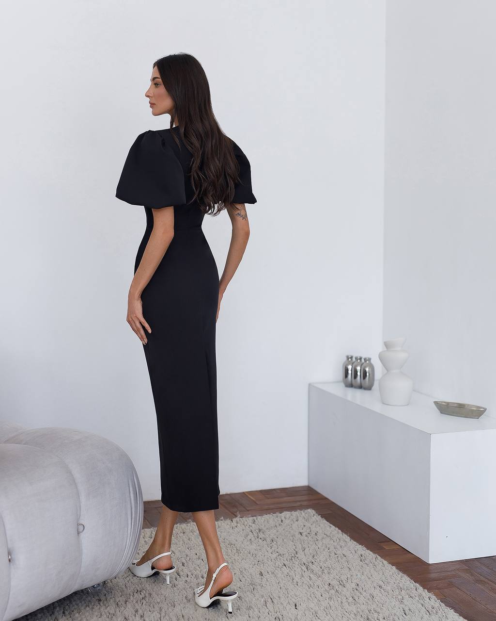 Woman in a black dress standing in a minimalistic room with a white wall and gray rug.