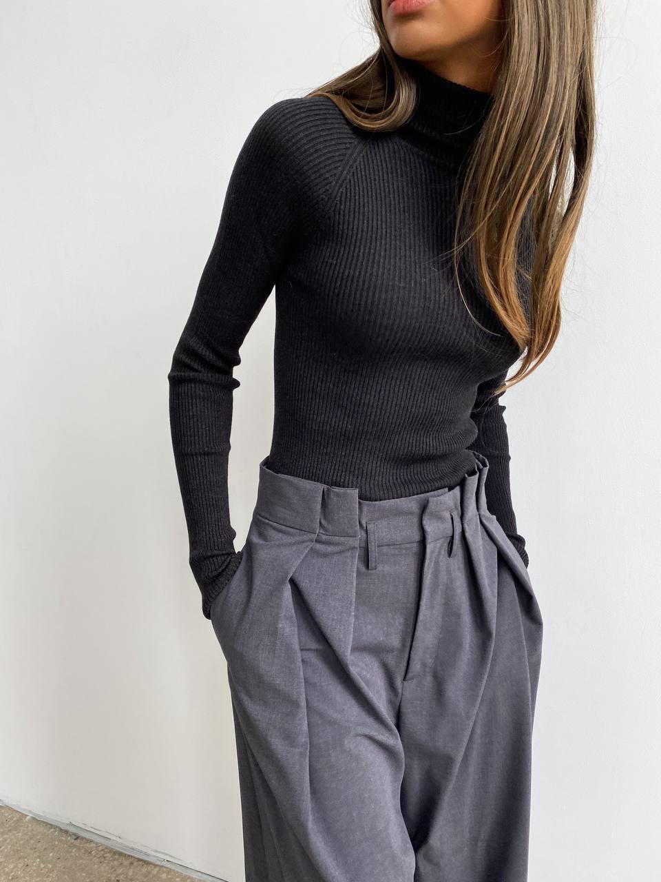 Person wearing a black turtleneck sweater and gray pleated pants against a white background