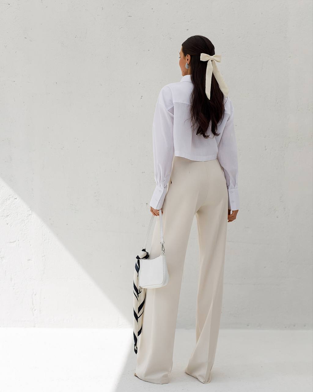 Person wearing a white shirt and beige pants with a black and white bag, standing against a light gray background