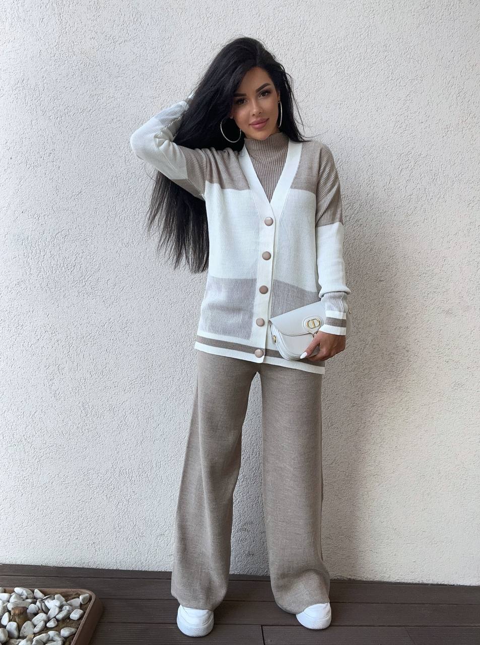 Woman wearing a beige and white cardigan with matching pants against a plain wall.