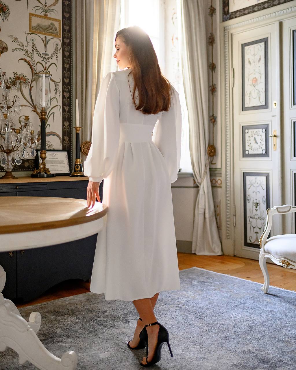 Woman in a white dress standing in an elegant room with decorative walls and furniture.