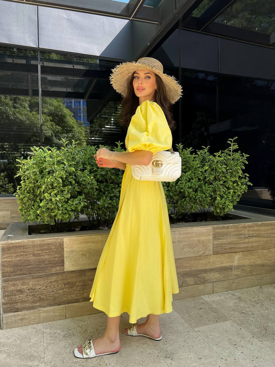 Woman in a yellow dress and hat standing outdoors with greenery and a building in the background