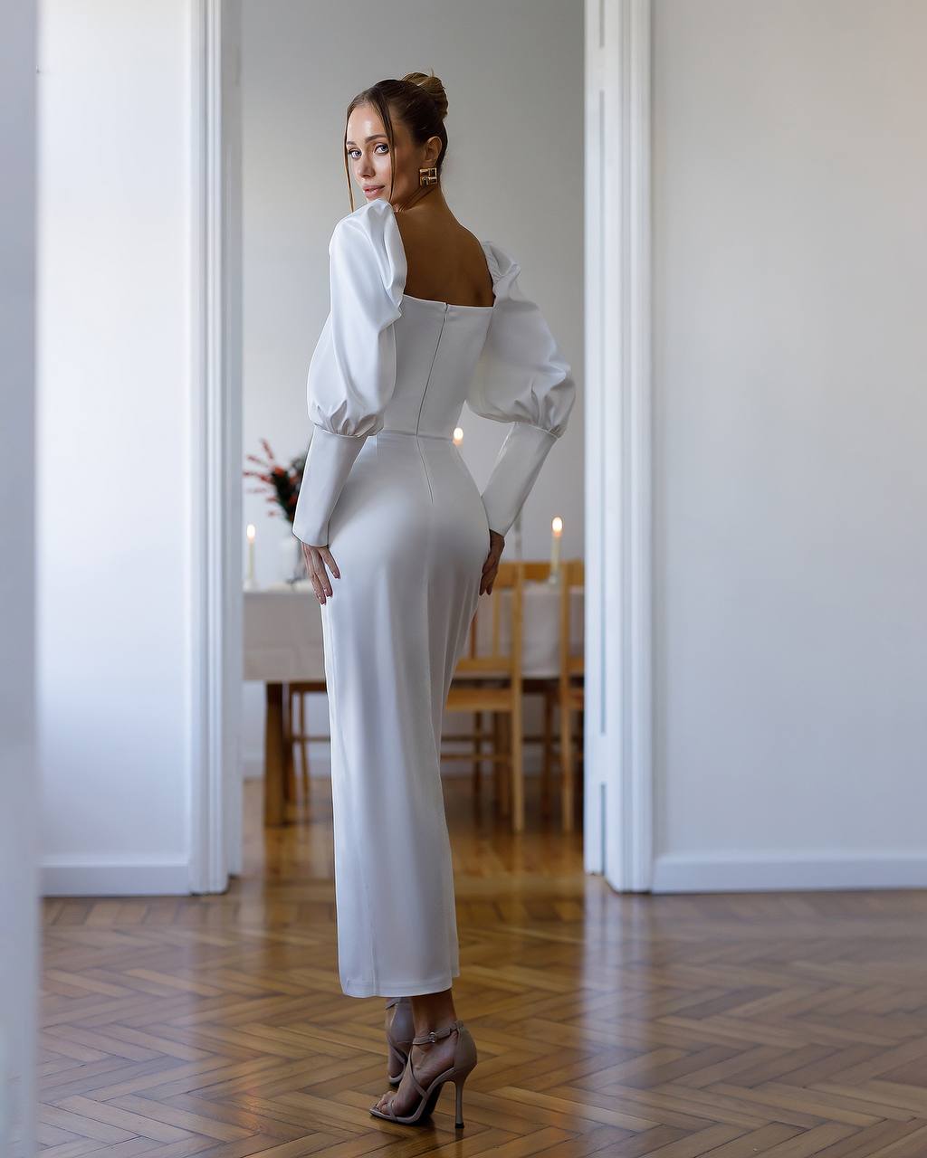 Woman in a white dress standing in a room with wooden flooring and white walls.