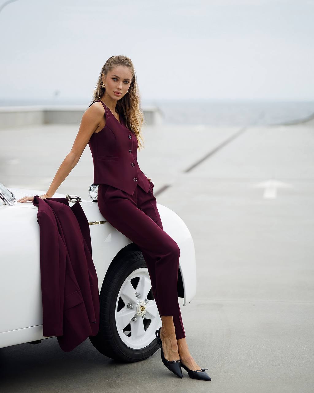 Woman in a burgundy outfit sitting on a white car in an indoor setting