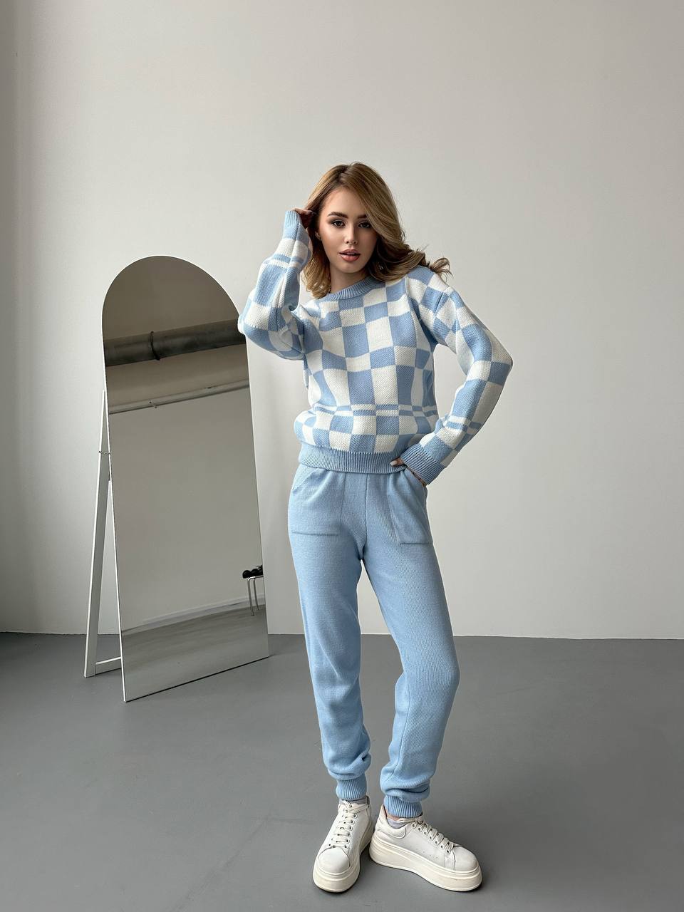 Woman wearing a blue and white checkered sweater and light blue pants, standing in a minimalistic room with a mirror.